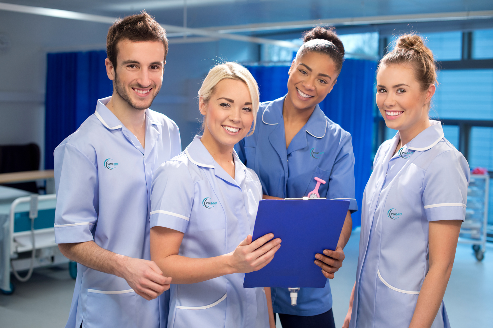 A diverse group of seven doctors, including men and women in white coats and scrubs, stand smiling in a bright medical facility. They all have stethoscopes around their necks.