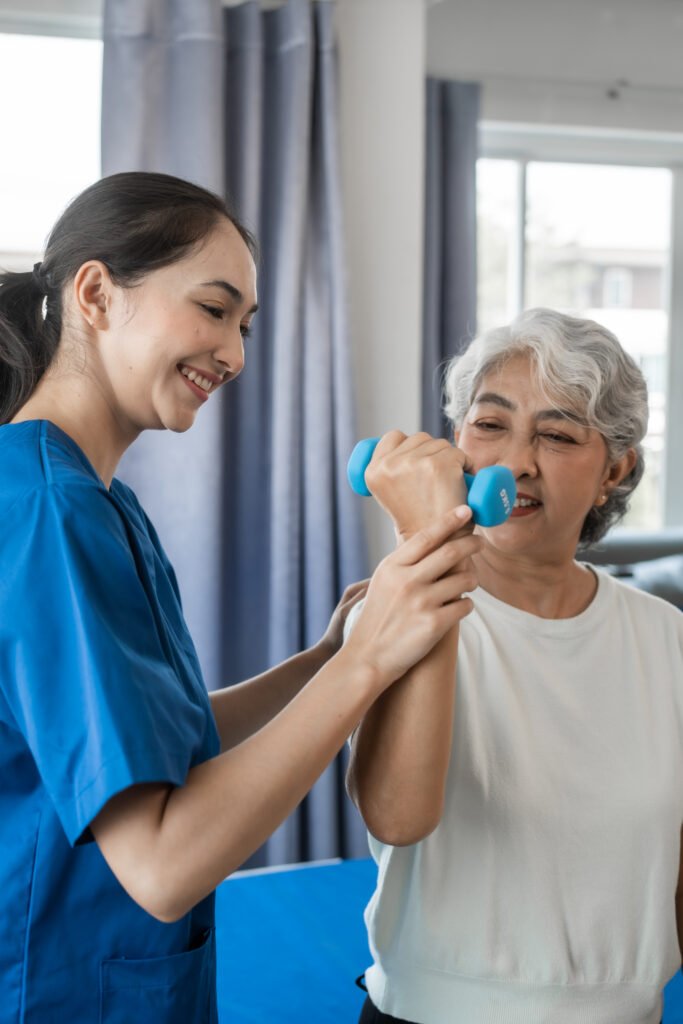 A young physiotherapist in a blue uniform assists a smiling senior mature Asian woman with grey hair in lifting a light blue dumbbell. They are indoors, with blurred window and curtains, suggesting a supportive exercise session.