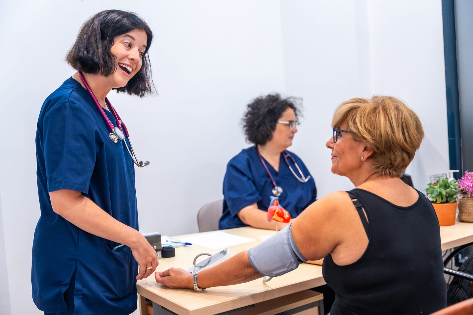 A healthcare worker in blue scrubs smiles while checking a womans blood pressure at the cardiology clinic. Another cardiologist in the background takes notes, ensuring all details are captured. A plant adds a touch of nature to the setting.