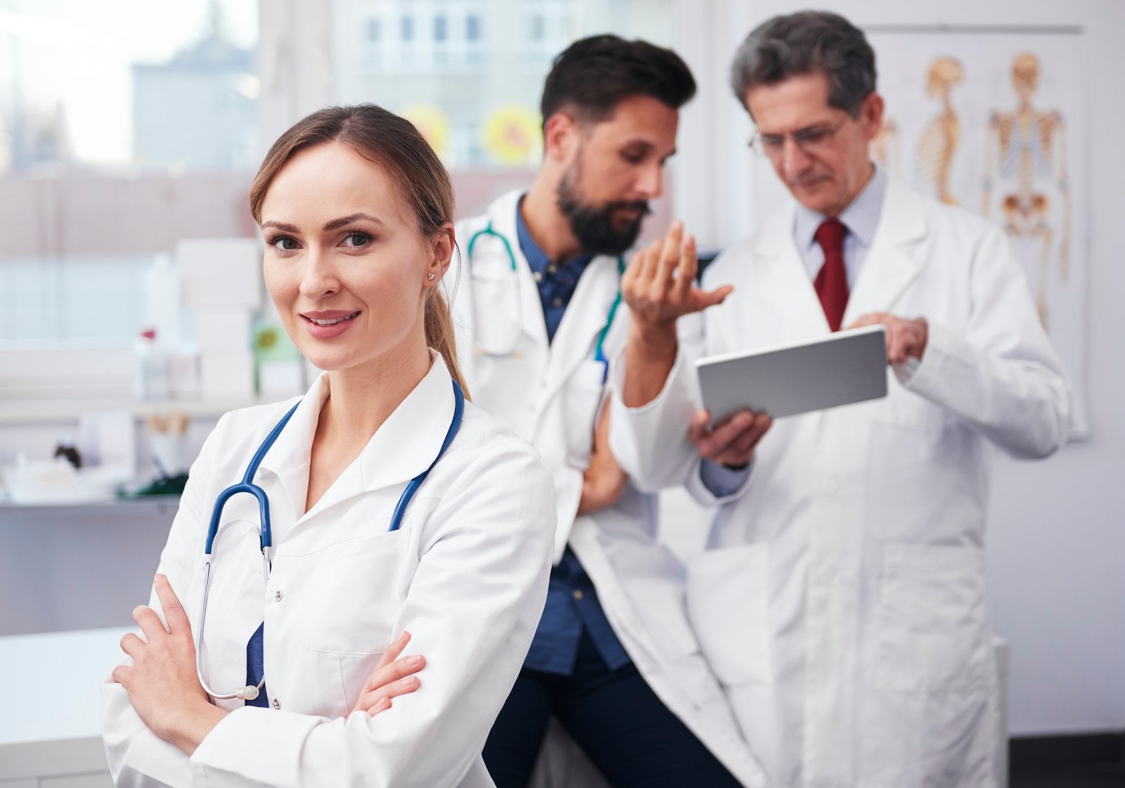 A portrait of a confident female doctor, stethoscope in hand, with arms crossed. In the background, two male doctors discuss consultations-with-doctors while referencing a tablet. All are in white lab coats within a clinical setting.