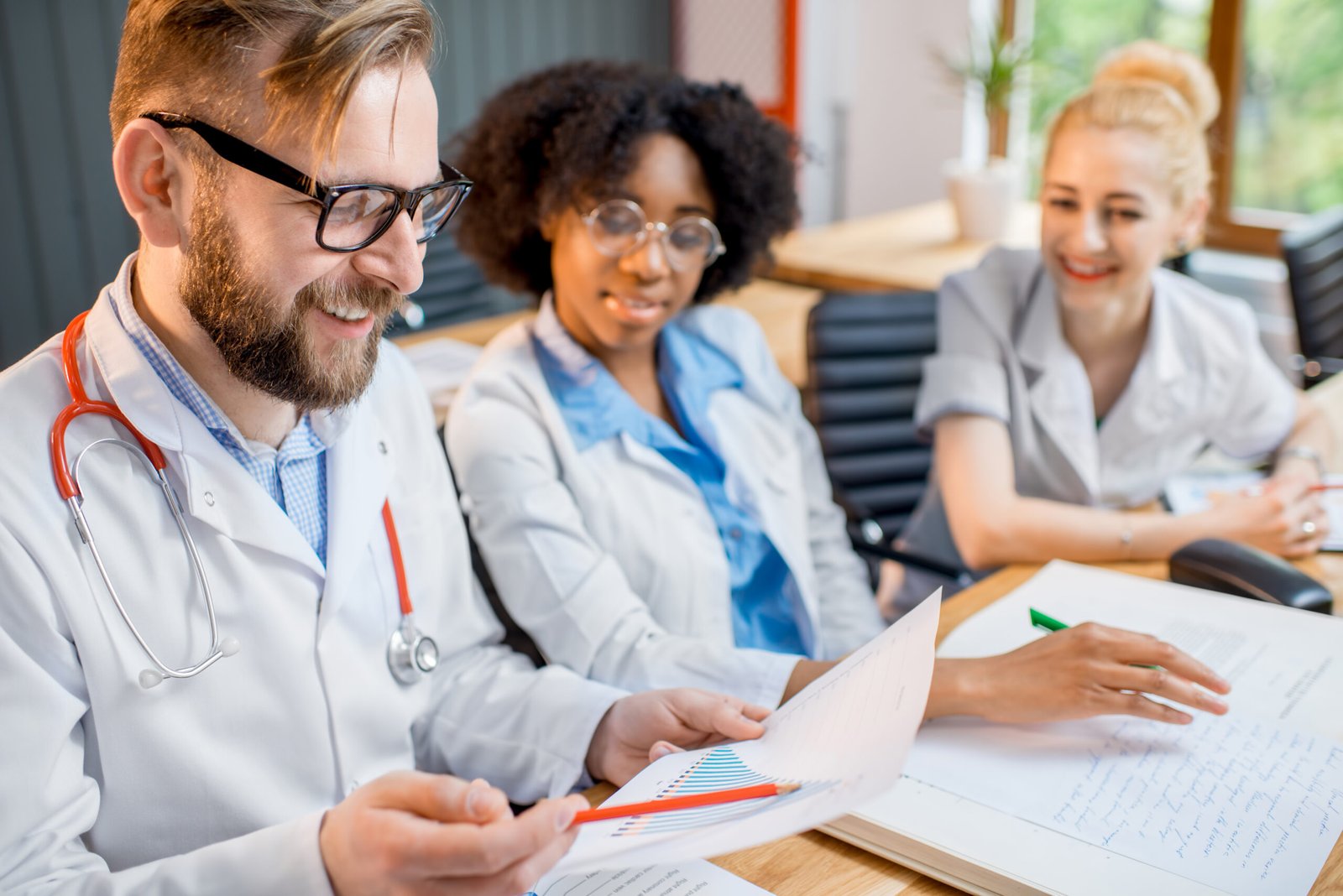 Three medical professionals in lab coats, resembling a group of medical students in a classroom, sit at a table reviewing documents and charts. A man with glasses holds a paper, while two women—one with curly hair and the other with a bun—look on and smile in the brightly lit room.