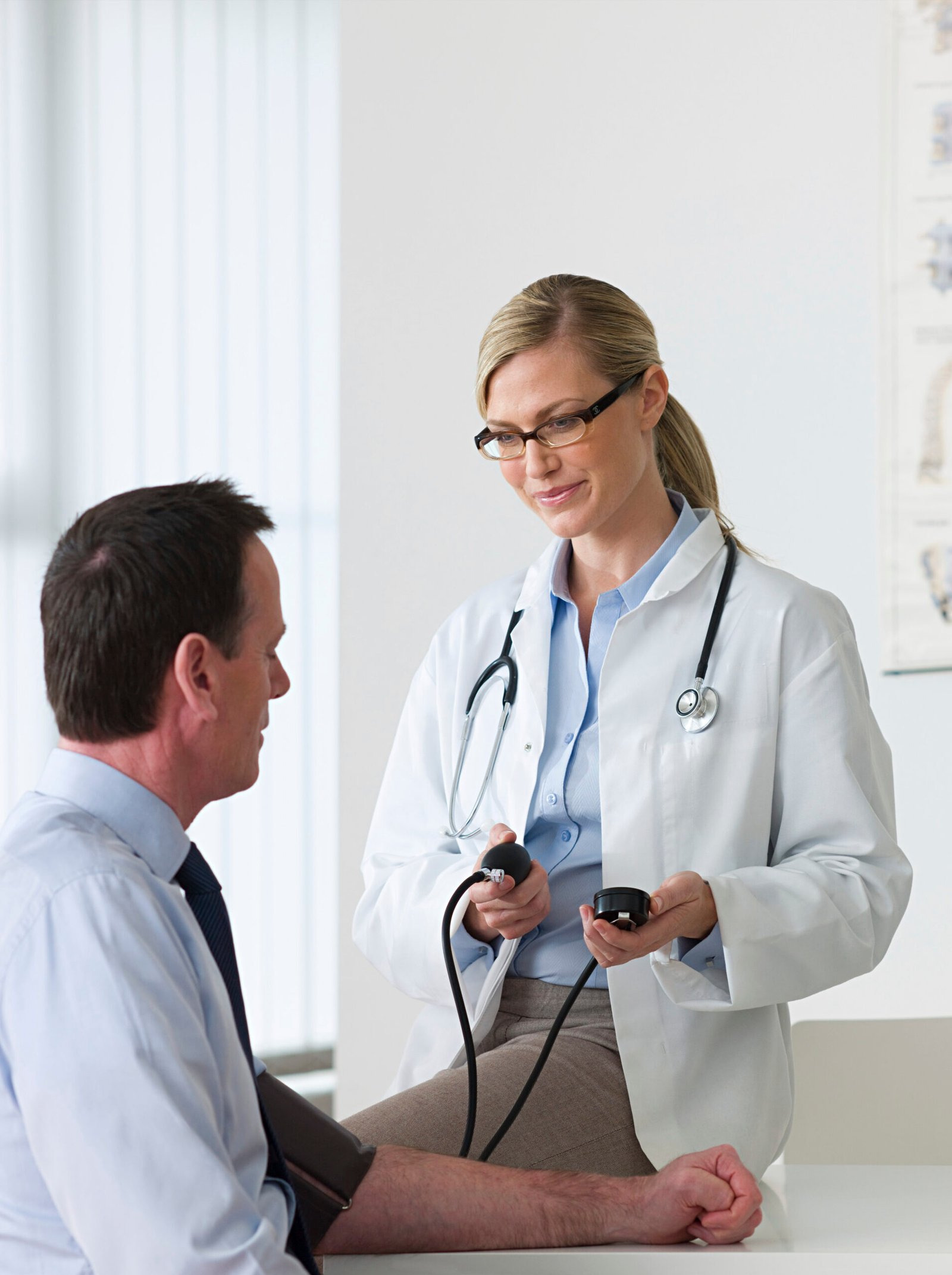 A doctor with glasses and a stethoscope, wearing a white coat, is conducting a check-up by measuring the blood pressure of a man in a blue shirt seated on an examination table. The room maintains its bright, clinical appearance.