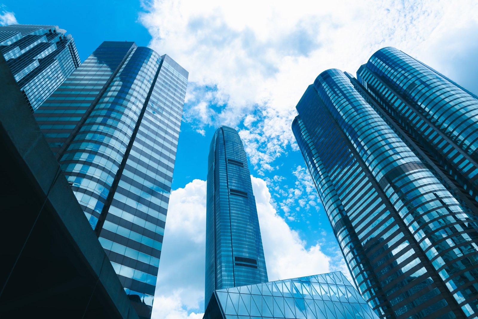 Tall, modern skyscrapers with reflective glass windows rise against a bright blue sky dotted with clouds. The sleek architecture of the skyscraper-building-in-Hong-Kong-city-view-in-blue-filter features cylindrical and rectangular shapes, creating a dynamic urban skyline.