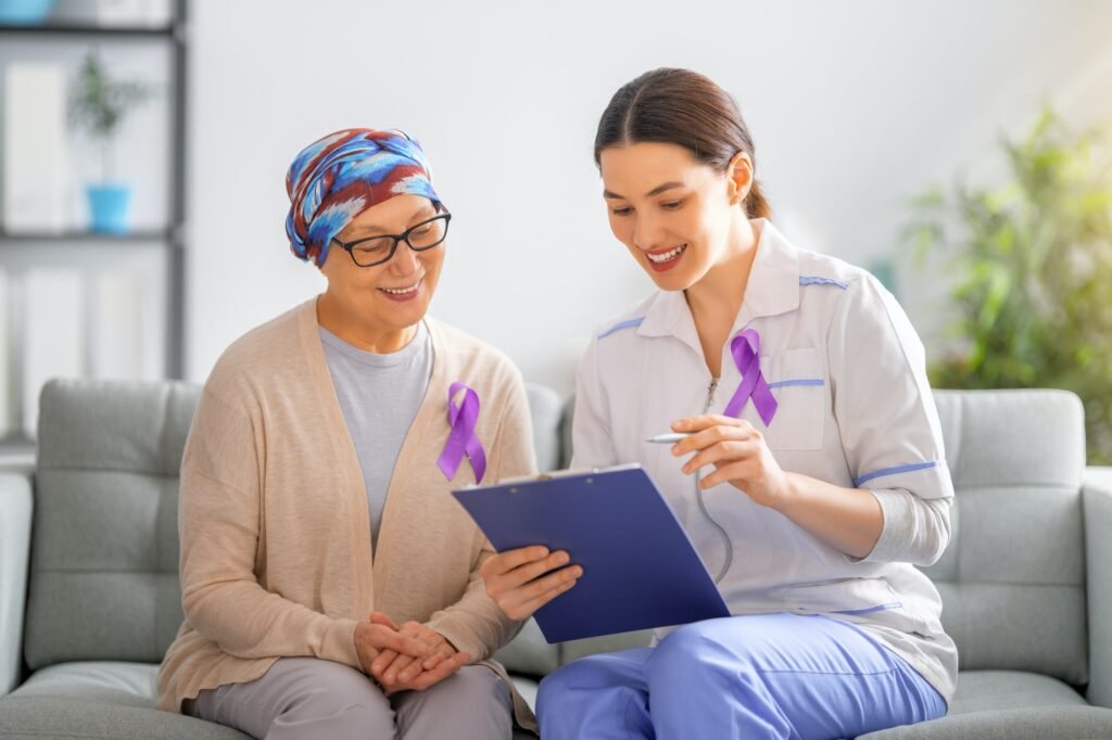 A woman in a headscarf and a nurse with a clipboard sit together on the couch, smiling. Purple ribbons on their clothing highlight their support for World Cancer Day. The softly blurred background features plants and shelves, adding to the serene atmosphere.